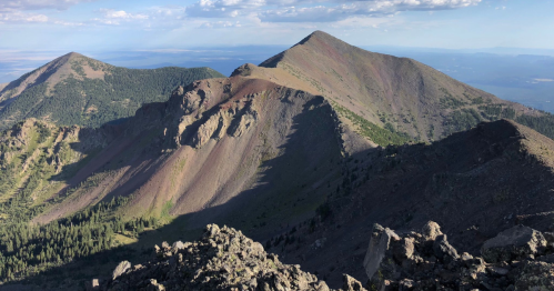 A panoramic view of rugged mountain peaks under a clear blue sky, with shadows highlighting the terrain.