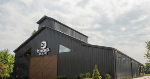 A large, modern distillery building with a dark exterior and a logo, set against a cloudy sky.