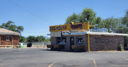 A small brick building with a bright yellow sign reading "Dog House," surrounded by trees and a clear blue sky.