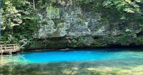 A serene blue pool surrounded by lush greenery and rocky cliffs, with a wooden walkway nearby.
