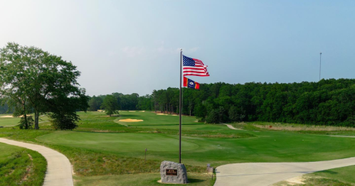 A golf course with a U.S. flag and a state flag flying, surrounded by trees and a clear sky.