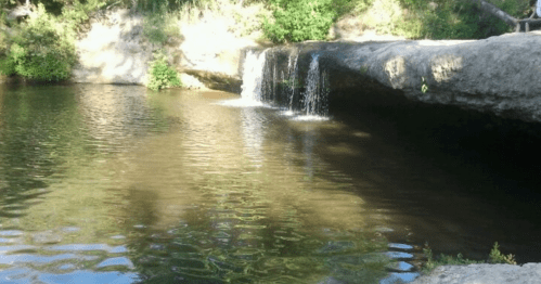 A serene waterfall cascading into a calm pool, surrounded by lush greenery and rocky terrain.