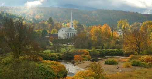 A scenic view of a church surrounded by colorful autumn trees and a flowing river under a cloudy sky.