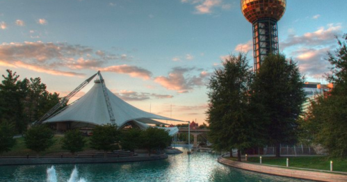 A scenic view of a park with a large tent structure, a tower, and a calm water feature under a colorful sky.