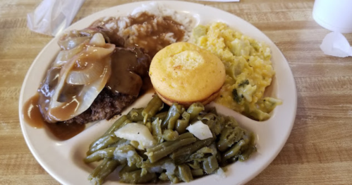 A plate of comfort food featuring meatloaf with gravy, mashed potatoes, green beans, cornbread, and cheesy vegetables.
