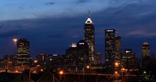 Indianapolis skyline at dusk, featuring tall buildings and a lit-up cityscape against a twilight sky.