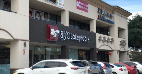 Exterior of a bakery café with a sign, surrounded by parked cars and other storefronts in a shopping plaza.