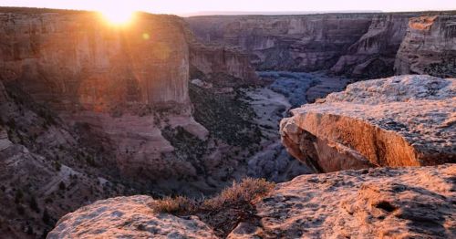 Sunrise over a canyon, casting warm light on rocky cliffs and a winding river below, surrounded by rugged terrain.