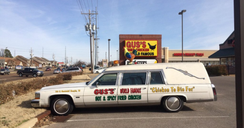 A unique hearse-style vehicle advertising Gus's World Famous Fried Chicken, featuring colorful text and a humorous slogan.