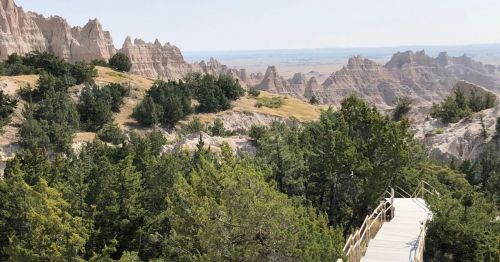 A wooden walkway winds through lush greenery, leading to rugged rock formations and a vast landscape in the distance.