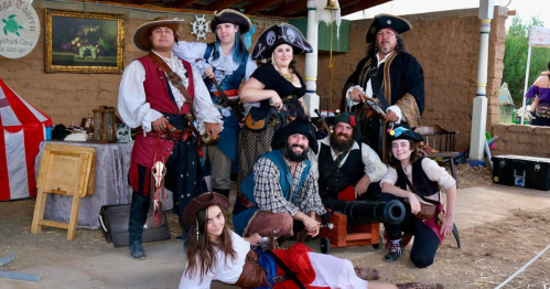 A group of eight people dressed as pirates, posing together in a rustic setting with props and costumes.