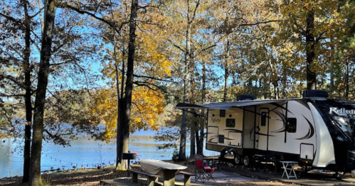 A camper parked by a serene lake surrounded by autumn trees with colorful leaves. A picnic table is nearby.