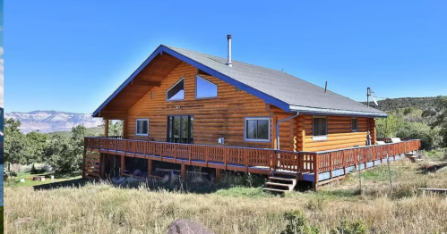 A wooden log cabin with a large deck, surrounded by grass and trees, under a clear blue sky.