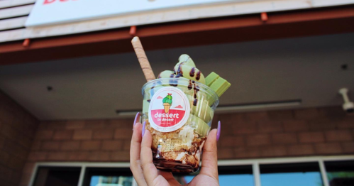 A hand holds a dessert cup with ice cream, chocolate drizzle, and a wafer, in front of a dessert shop sign.