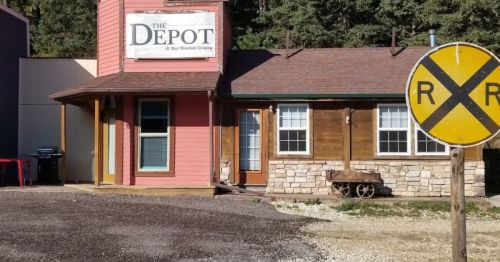 A quaint building labeled "The Depot" with a railroad sign nearby, surrounded by trees and gravel.