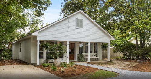 A charming white house with a front porch, surrounded by greenery and a gravel pathway.