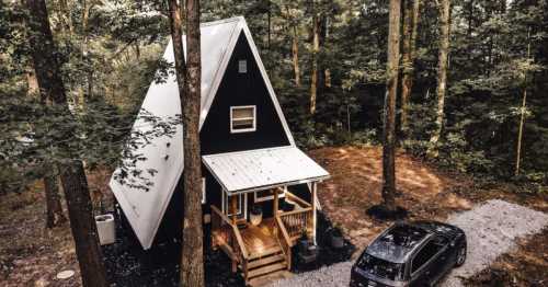 Aerial view of a modern A-frame cabin in a wooded area, with a gravel driveway and a parked car.