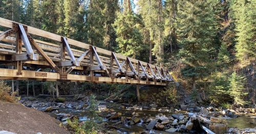 A wooden bridge spans a creek, surrounded by tall trees and rocky terrain in a forested area.