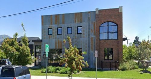 A modern building with a mix of metal and brick exterior, featuring large windows and surrounding greenery.