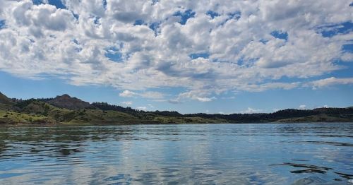 A serene lake reflecting fluffy clouds and green hills under a bright blue sky.