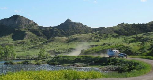 A scenic landscape with rolling green hills, a lake, and a vehicle driving along a dirt road.