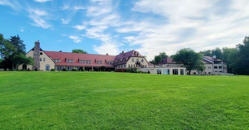 A large, elegant building with a red roof sits on a green lawn under a blue sky with scattered clouds.