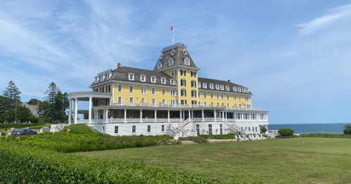 A large yellow historic hotel with a clock tower, surrounded by green lawns and overlooking the ocean under a blue sky.