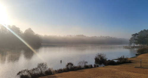A serene lake at sunrise, with mist rising from the water and trees lining the shore under a clear blue sky.