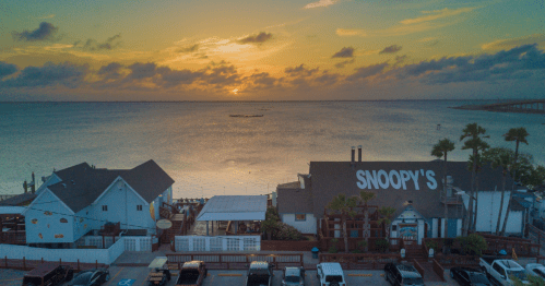 A sunset over the water with a restaurant named "Snoopy's" in the foreground and palm trees lining the shore.
