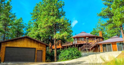 A round wooden house with a deck, surrounded by tall pine trees, next to a garage on a sunny day.