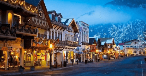 Charming street lined with festive buildings, adorned with lights, set against a misty mountain backdrop at dusk.