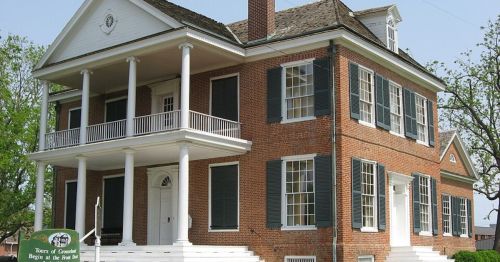 Historic brick house with white columns, green shutters, and a porch, surrounded by trees and a sign in front.