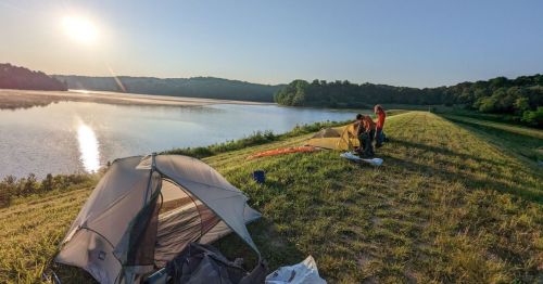 Two people set up tents by a serene lake at sunrise, with lush greenery in the background.