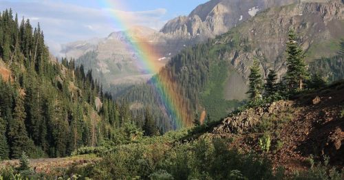 A vibrant rainbow arcs over a lush green valley surrounded by mountains and pine trees.