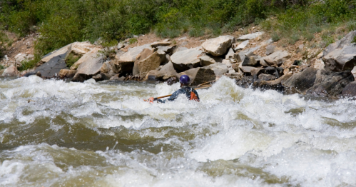 A kayaker navigates through turbulent whitewater rapids surrounded by rocky banks and greenery.
