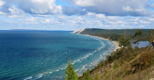 A scenic view of a coastline with blue waters, sandy beach, and green hills under a partly cloudy sky.
