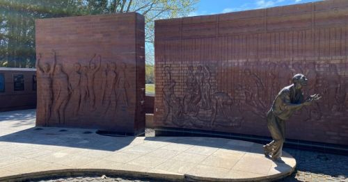 A bronze sculpture beside a brick wall with relief figures, set in a park under a clear blue sky.