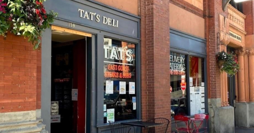 Exterior of Tat's Deli, featuring a sign and outdoor seating, with brick architecture and hanging flower baskets.