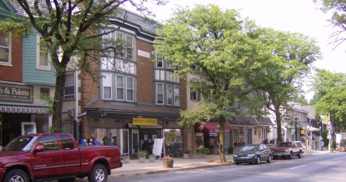 A quaint street scene featuring shops, trees, and parked cars in a small town.