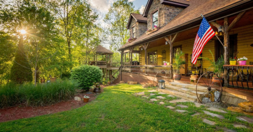 A charming house with a porch, American flag, and lush greenery, surrounded by trees and a sunny sky.