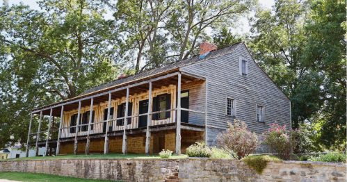 Historic wooden house with a porch, surrounded by trees and greenery.
