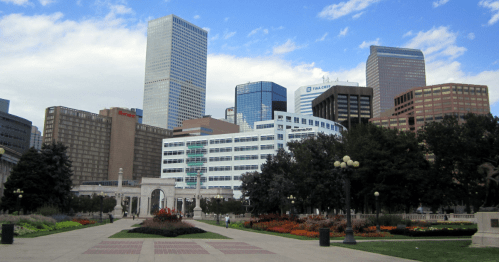 City skyline with modern buildings and green park space, featuring flowers and trees under a partly cloudy sky.