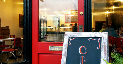A red door with an "Open" sign leads into a cozy café with tables and a display case visible inside.