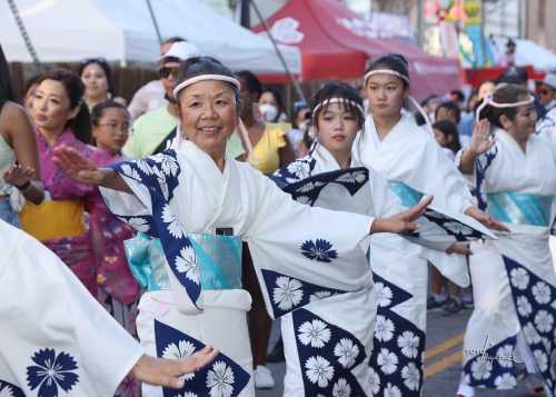 A group of women in traditional Japanese attire perform a dance at a festival, surrounded by onlookers.