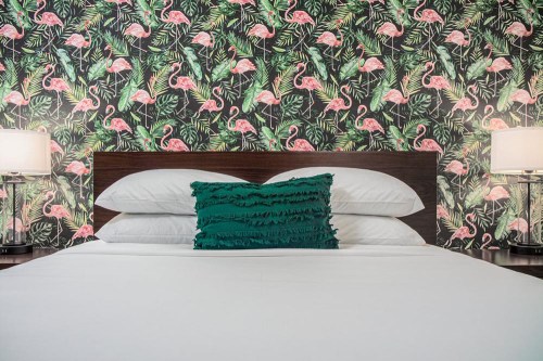 A bed with white linens and a green ruffled pillow, set against a vibrant flamingo-patterned wallpaper.