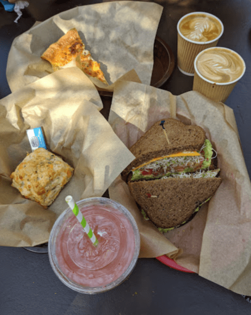 A table with a sandwich, a biscuit, a slice of pizza, and two cups of coffee with latte art, alongside a pink drink.