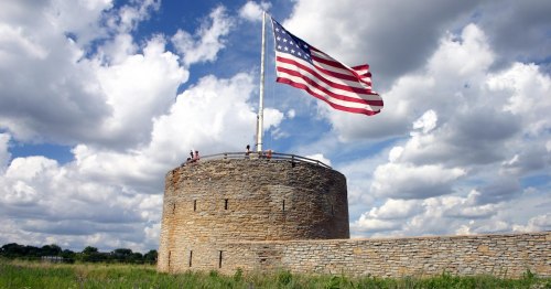 A stone round tower with an American flag flying, set against a backdrop of fluffy clouds and blue sky.