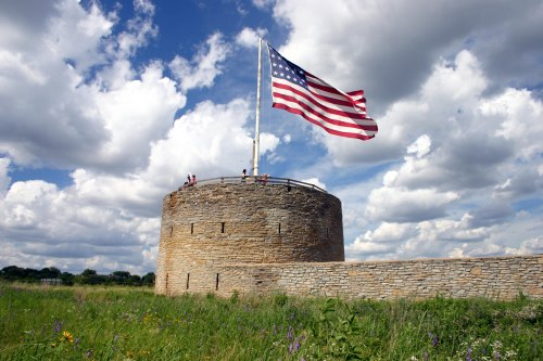 A stone tower with a large American flag, surrounded by green grass and under a cloudy blue sky.