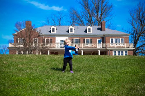 A child in a blue jacket plays in a grassy field with a large brick house in the background under a clear blue sky.