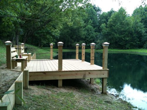 A wooden dock with posts beside a calm pond, surrounded by lush green trees.